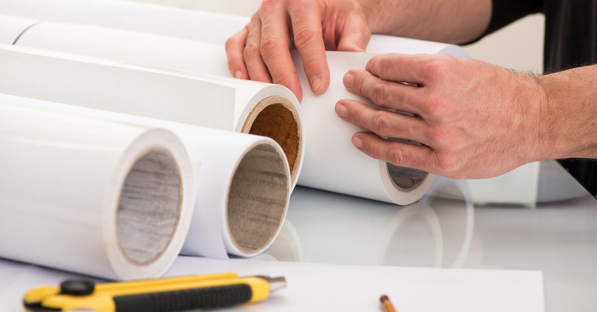 A person unfolds a large paper roll for a plotter printer. There is a box opener and measuring tape on the table.