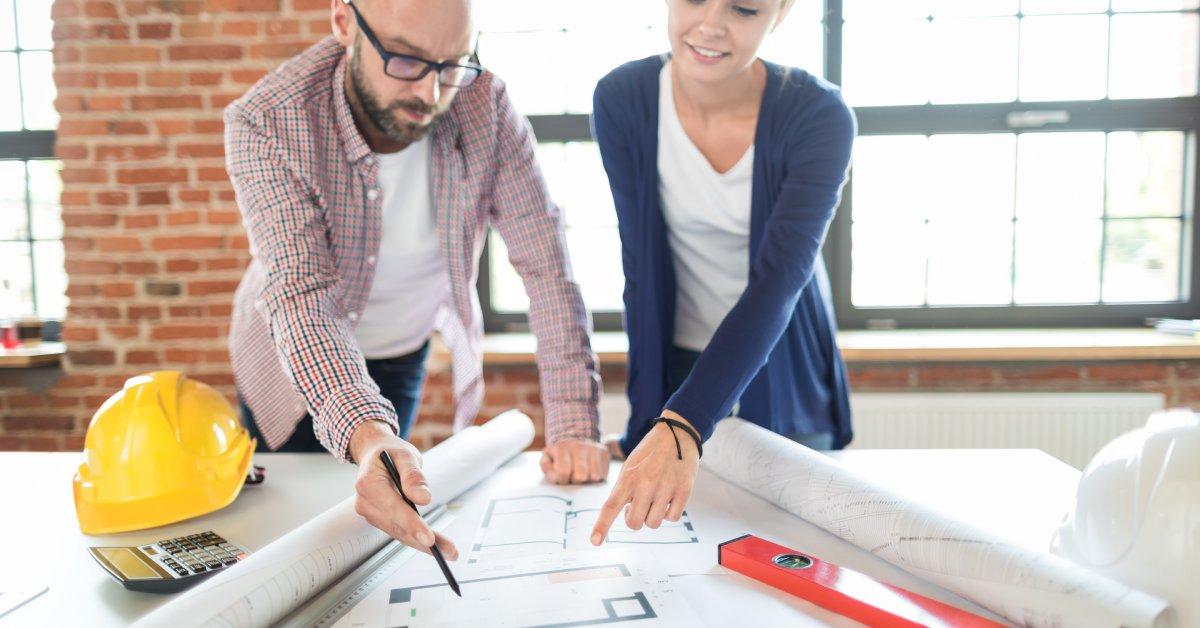 Two professional architects spread large blueprints for a construction site at their office table and take notes.