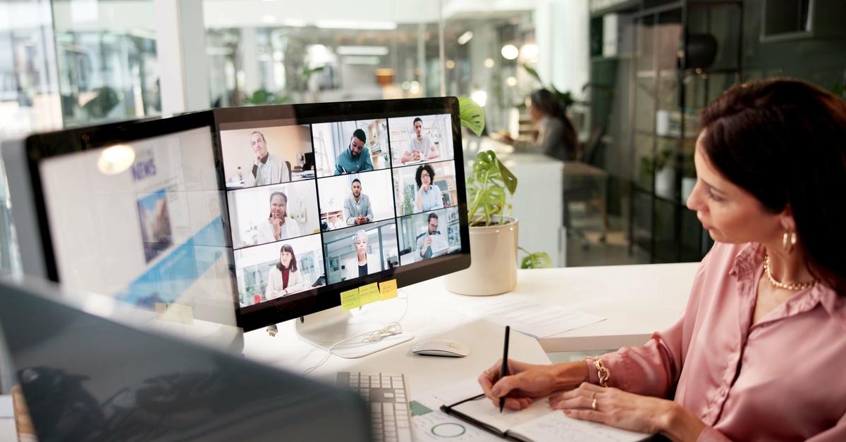 A businesswoman sits at her office desk and writes notes during a video call with nine other professionals.