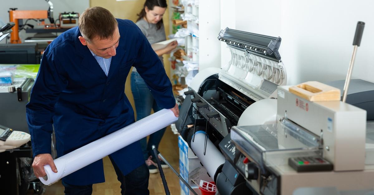 A worker loads a new roll of paper into a wide-format plotter. Another worker in the background organizes documents.