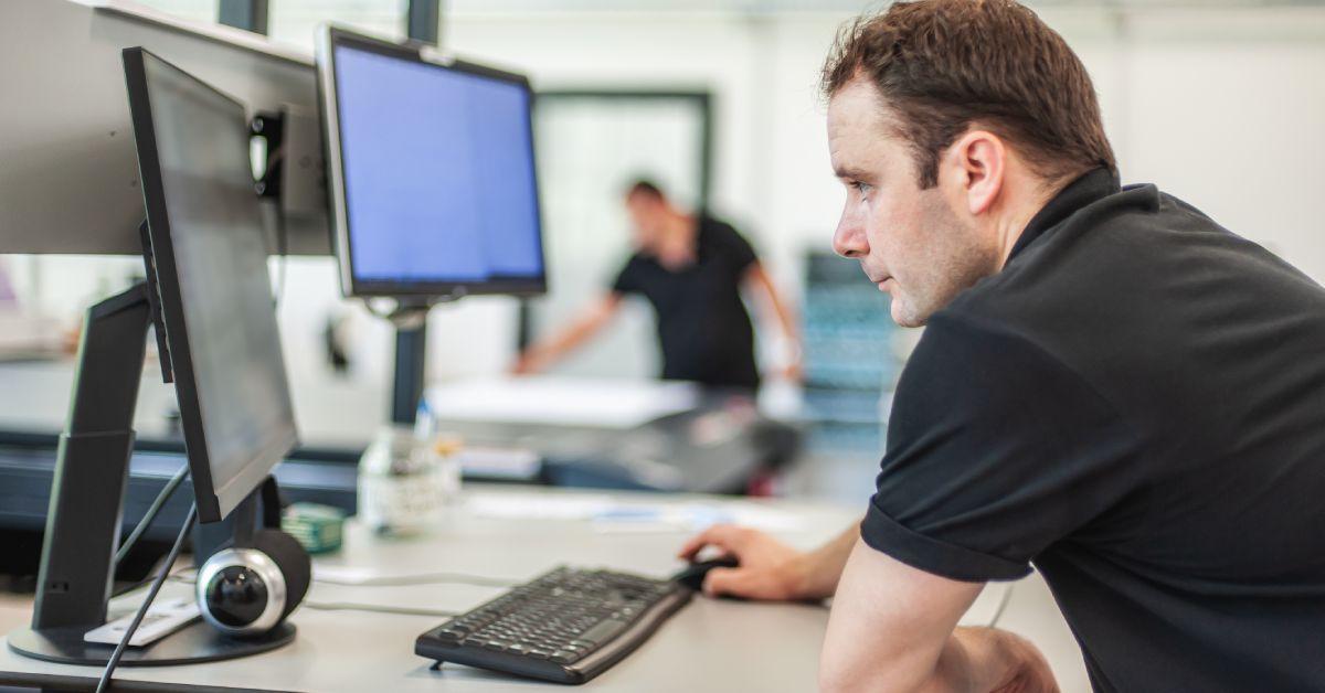 A technician works on a computer to assess project details in a printing shop. Another worker is in the background.