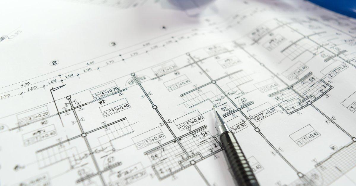A close-up of an engineering blueprint layout with detailed measurements. A black pencil rests on the material.