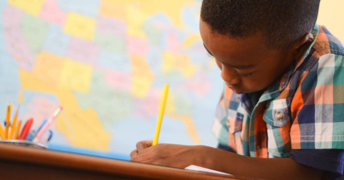 A young Black student uses a writing utensil at his desk, and a map of the United States is visible in the background.