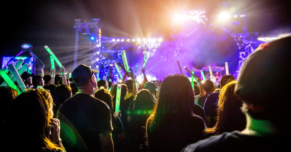A crowd of people holding glowing light sticks faces a large stage with bright lighting and purple effects.