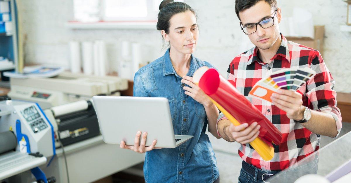A man looks at a color palette while holding rolls of material, and a woman with a laptop stands beside him.