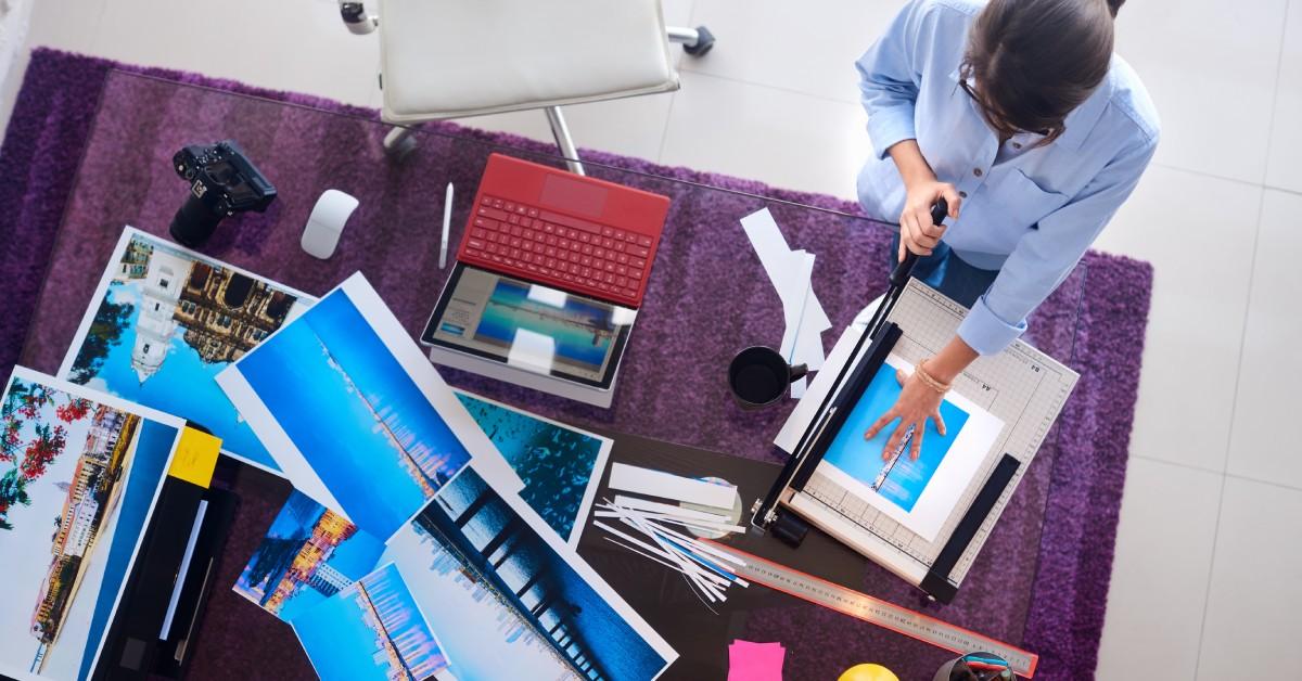 Overhead view of a desk with a person trimming photos using a paper cutter, surrounded by prints, a camera, and a laptop.