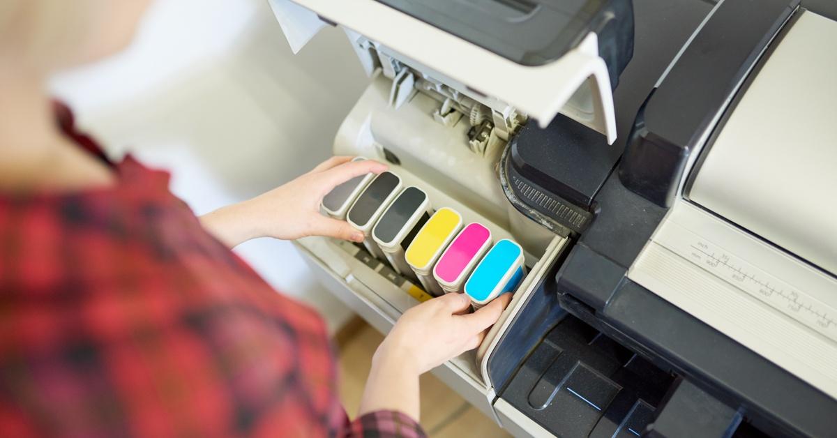 A person loads ink cartridges into a large-format printer, viewed from above, with printer housing and controls visible.