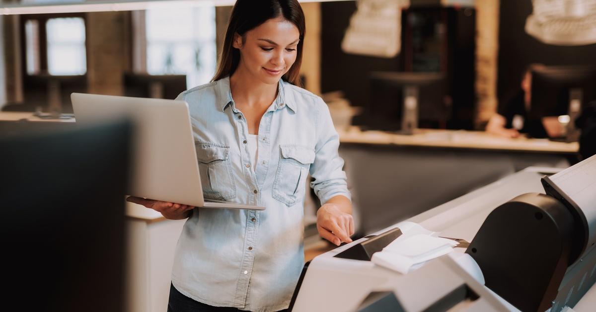A woman holds a laptop while operating a large printer in a print shop, checking the controls as paper feeds through.