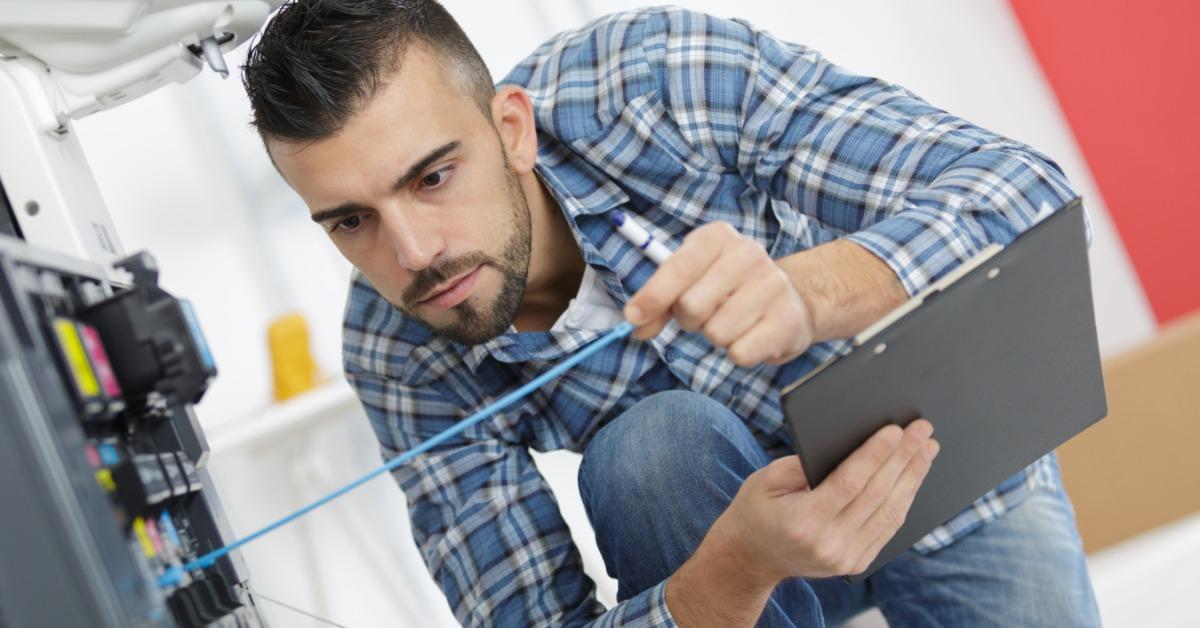 A man in a blue flannel shirt checks the ink levels of a printer with a blue stick while holding a black clipboard.