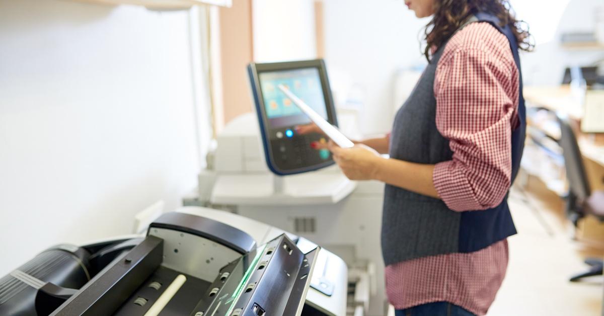 A woman wearing glasses stands in front of a plotter printer, pressing buttons on the control panel beside her.