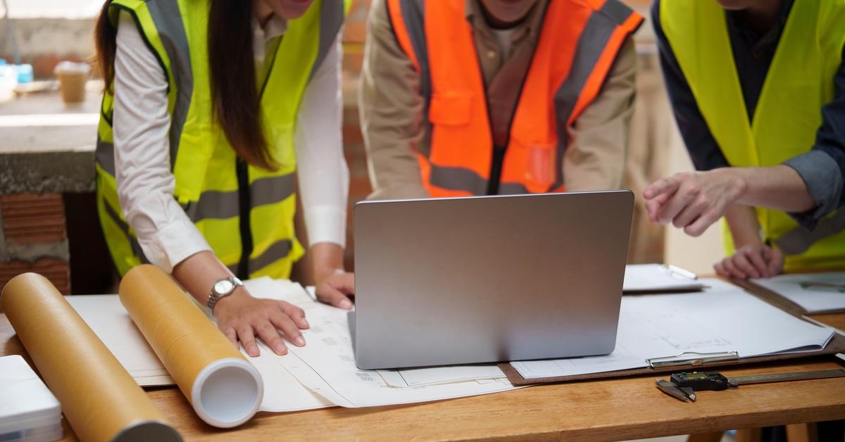 People reviewing architectural drawings and digital plans on a laptop while gathered around a wooden table indoors.