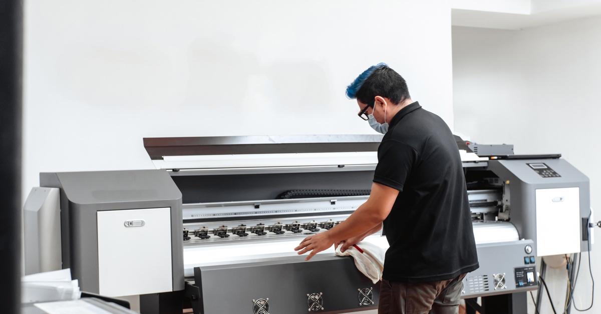 A person wearing a mask cleans a large-format printing machine inside a workspace with equipment and tools visible.