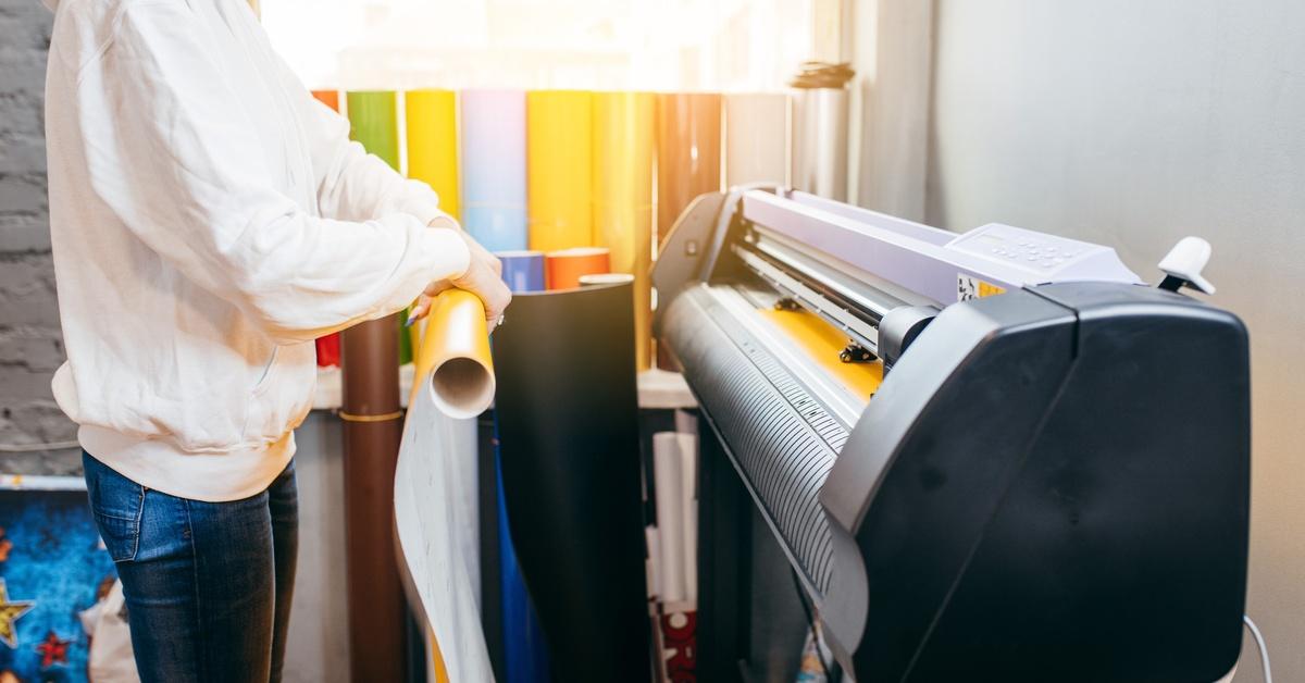 A person holding a sheet of yellow printed material near a large-format machine in a production setting.