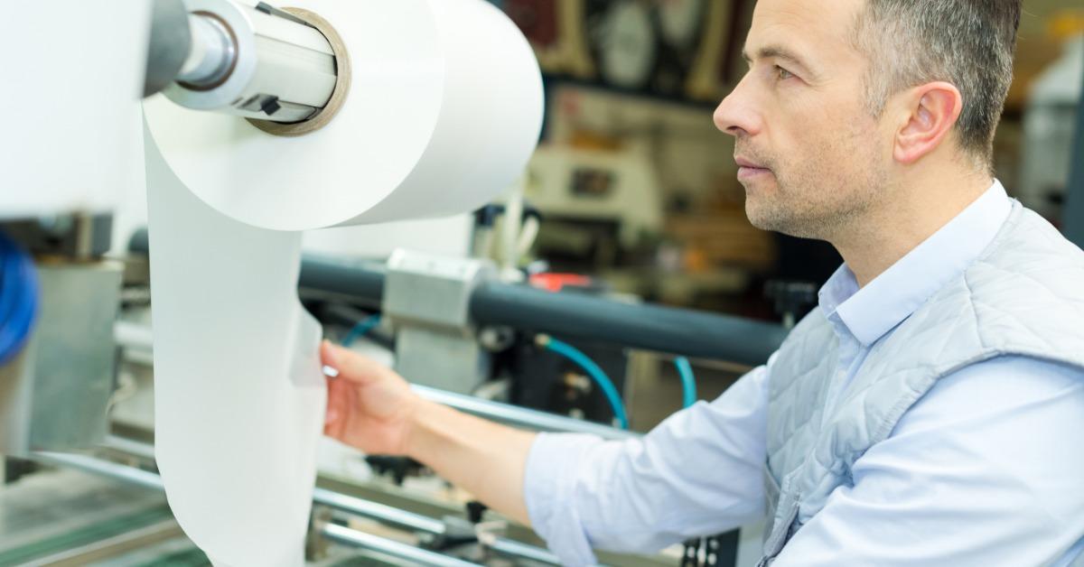 A man in a light shirt holds rolled white paper beside printing machinery inside an industrial production facility.
