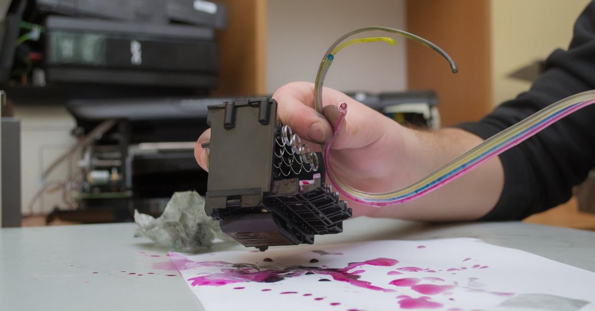 A close-up of a person refilling a printer's ink tank with a bottle of colored ink next to an open compartment.